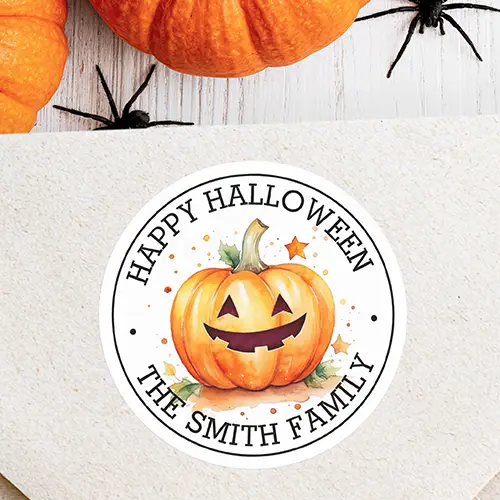 Circular white sticker featuring a smiling watercolor style pumpkin Jack-O'-Lantern illustration encircled by curved black typewriter text reading Happy Halloween along the top and custom name text along the bottom.
