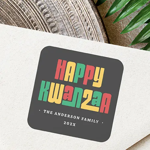Square self-adhesive label featuring red, green, and gold stacked Happy Kwanzaa typography on dark grey, with customizable white text at the bottom.