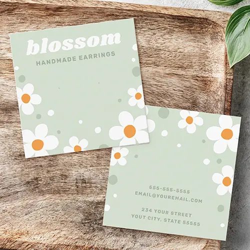 Two-sided square earring packaging card showing white daisy illustrations on a sage green background with custom business headline text and reverse contact info lines.