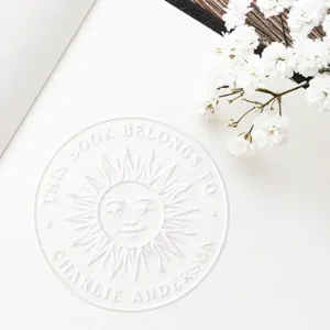 Boho sun character bookplate library book embosser A circular embossed white seal displaying a central celestial sun face surrounded by radial rays, with raised text reading This Book Belongs To and custom name or text along the border