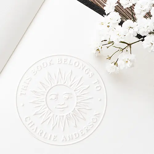A circular embossed white seal displaying a central celestial sun face surrounded by radial rays, with raised text reading This Book Belongs To and custom name or text along the border