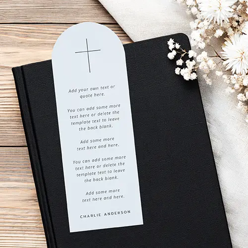 Arched paper bookmark in soft light blue featuring a dark cross icon at the top, italicized body text, and a dark uppercase name at the bottom.