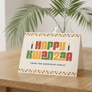 Horizontal folded greeting card with top and bottom geometric triangle borders in green, yellow, and red on a cream background, featuring Happy Kwanzaa block typography, candle icons, and a custom family name line. Customizable photo and text inside.
