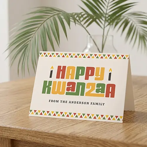 Horizontal folded greeting card with top and bottom geometric triangle borders in green, yellow, and red on a cream background, featuring Happy Kwanzaa block typography, candle icons, and a custom family name line. Customizable photo and text inside.