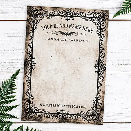A rectangular double-sided earring holder card featuring an ornate black filigree border on aged parchment paper with custom business text and a distressed dark back side.